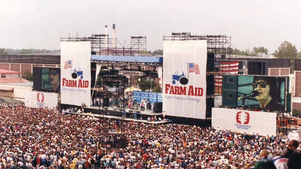 Packed outdoor concert venue with large crowd watching a performer on stage with giant banners by the stage