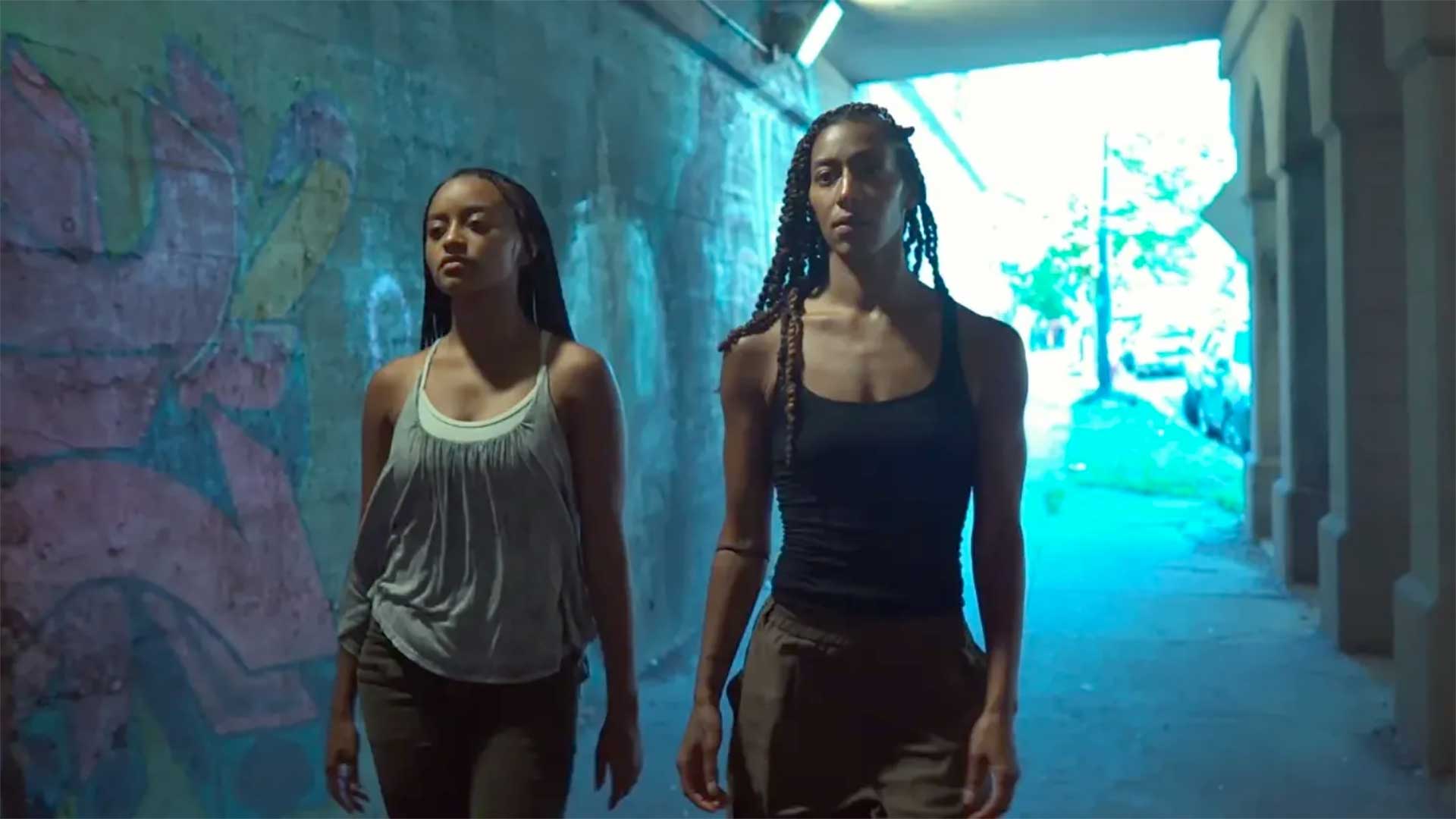 Two women walk through a dimly lit passageway under a viaduct, intently focused on what lies ahead