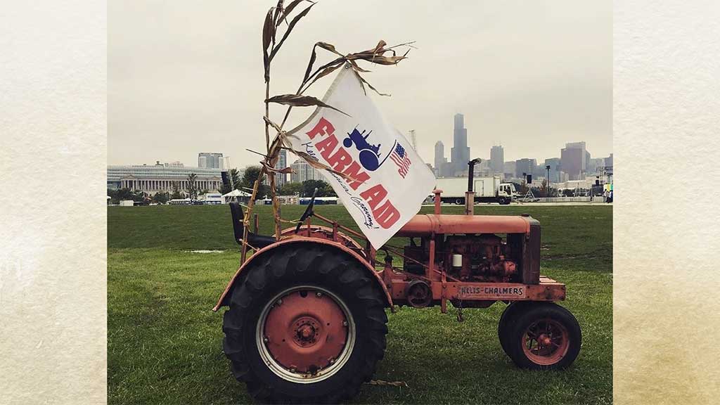 historic photo of a red tractor with farm aid flag and cornstalk on a greenspace with city skyline (possibly Chicago) in the background