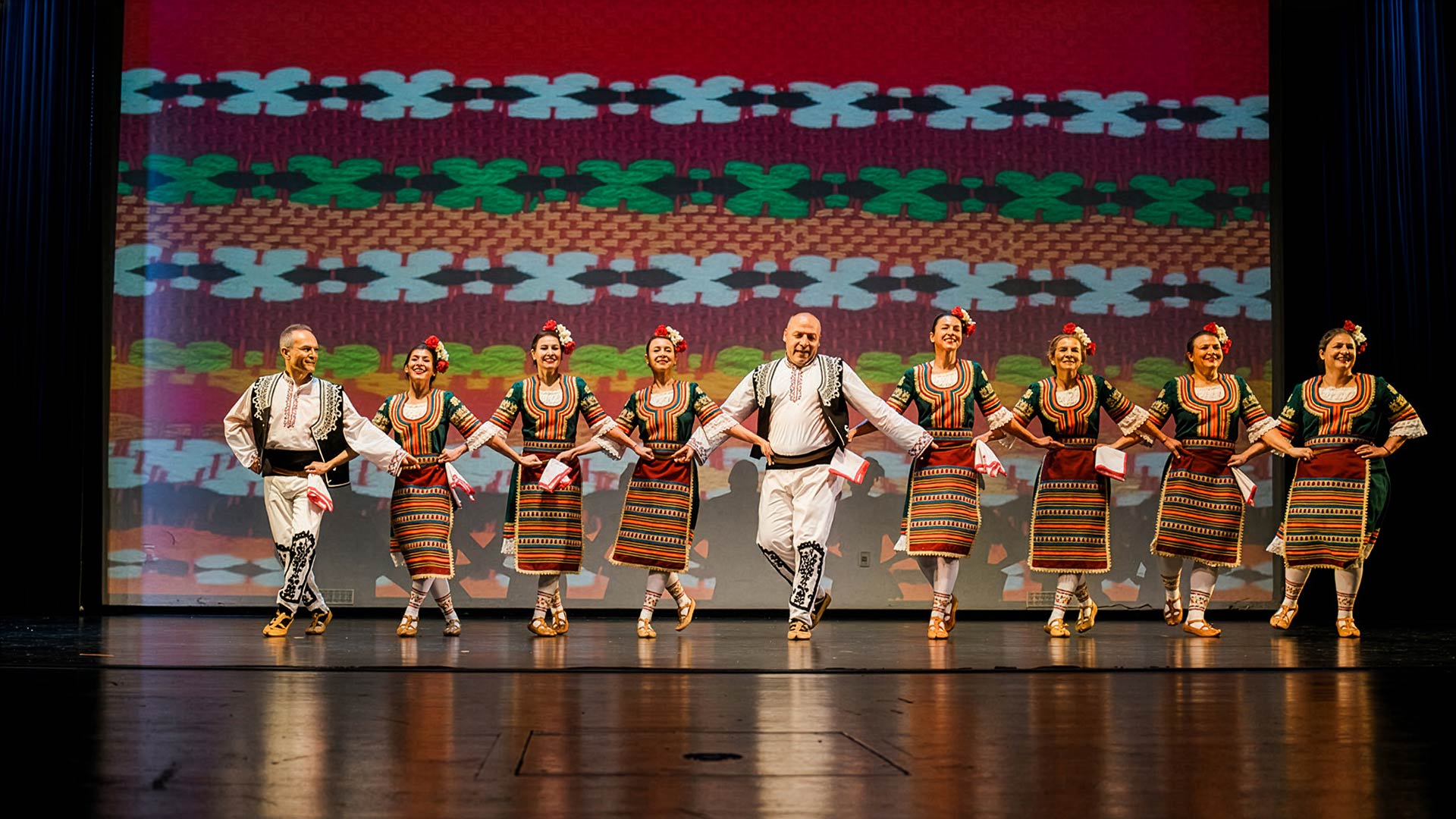 9 dancers wearing traditional bulgarian costumes in a line on an auditorium stage in front of background showing up a closeup of the texture of the women