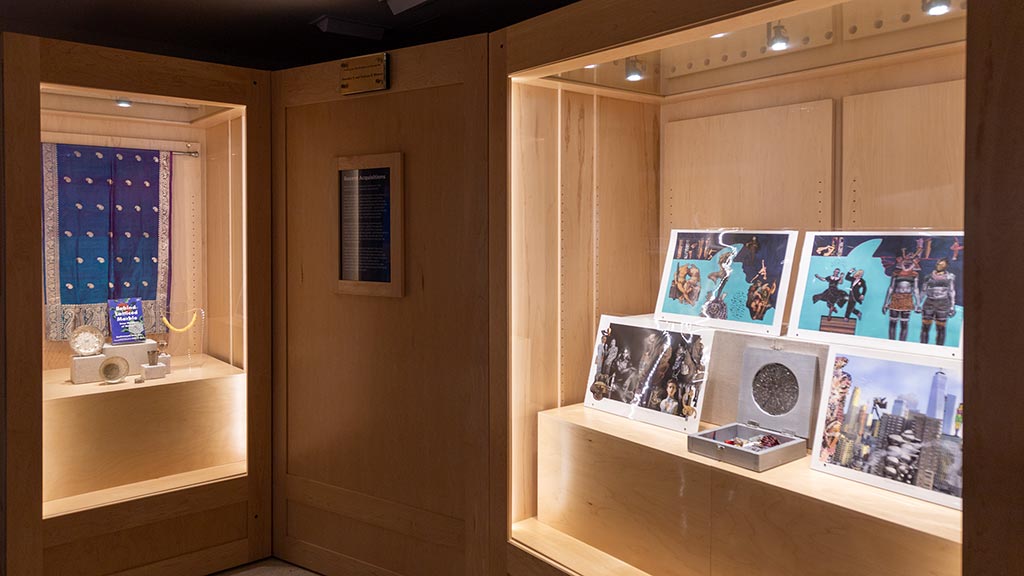 Two cases of objects featuring a blue fabric panel with book, smaller gold objects, and 4 larger collage-based art prints.