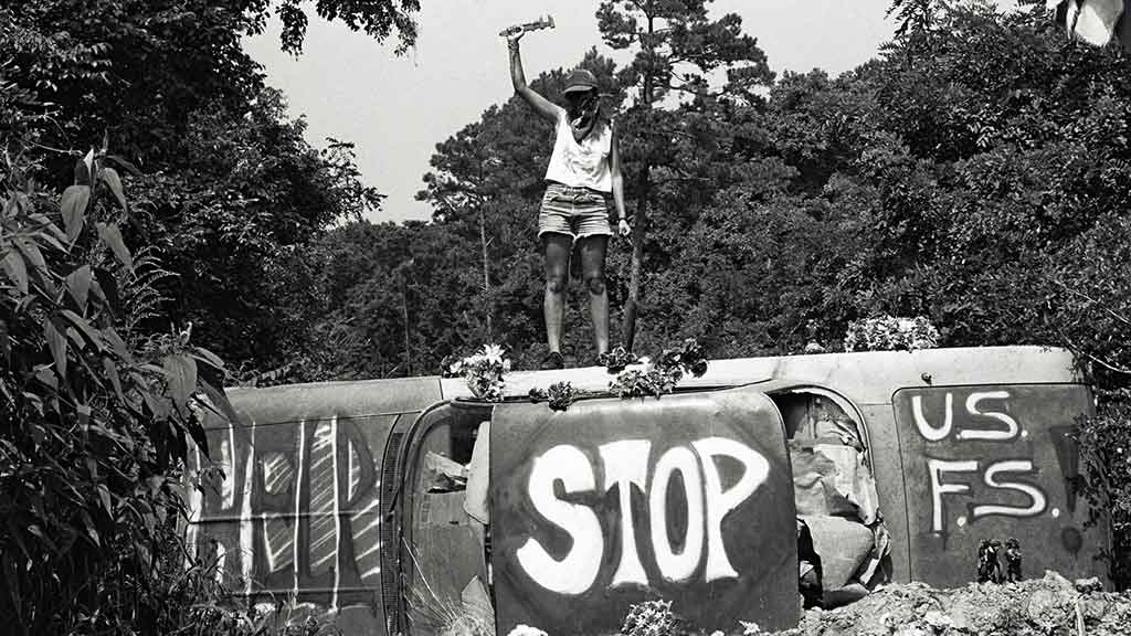 Greyscale picture of an activist standing on a tilted car with the words "Help" "Stop" and "US FS" painted on the car