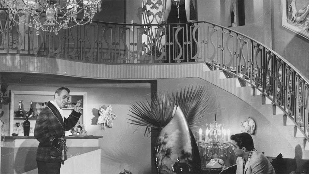 black and white film still of a lavishly decorated house with sweeping ornate staircase and two men in the foreground